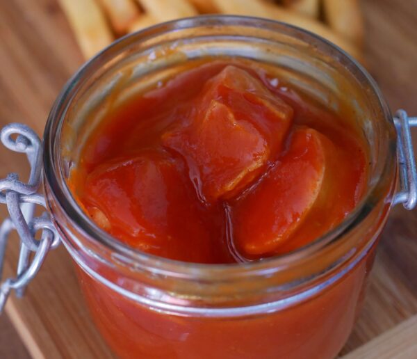 Ästhetische Draufsicht auf ein offenes Glas Berliner Currywurst, saftige Wurststücke in kräftigen roten Ketchup, vollständiger Glasrand ohne Deckel sichtbar.